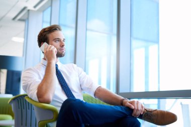 Yakışıklı genç bir işadamının geniş pencereli modern bir ofis alanında telefonla konuşurken ve dışarı bakarken düşük açılı görüntüsü.