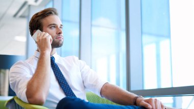 Parlak, modern bir ofis alanında rahat bir şekilde oturan yakışıklı bir iş adamının düşük açılı görüntüsü, telefonda konuşurken büyük pencerelerden dışarı bakıyor.