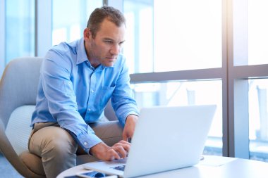 Büyük pencereli, parlak, modern bir ofiste yakışıklı bir işadamı, kahve masasında oturuyor ve bilgisayarındaki bazı bilgilere bakıyor.