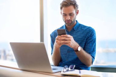 Modern ofiste önünde dizüstü bilgisayarı açık genç bir adam ve arkasında cep telefonuna mesaj yazan büyük bir pencere.