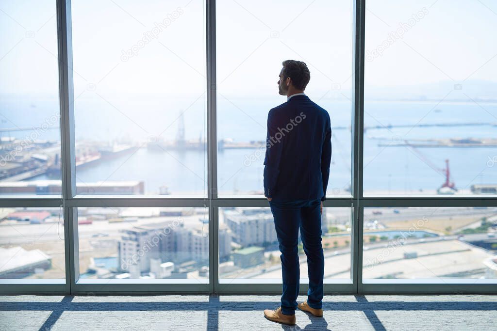 Full length rearview of a young stylish businessman standing at the large windows of a high office, looking at the view of a harbour below