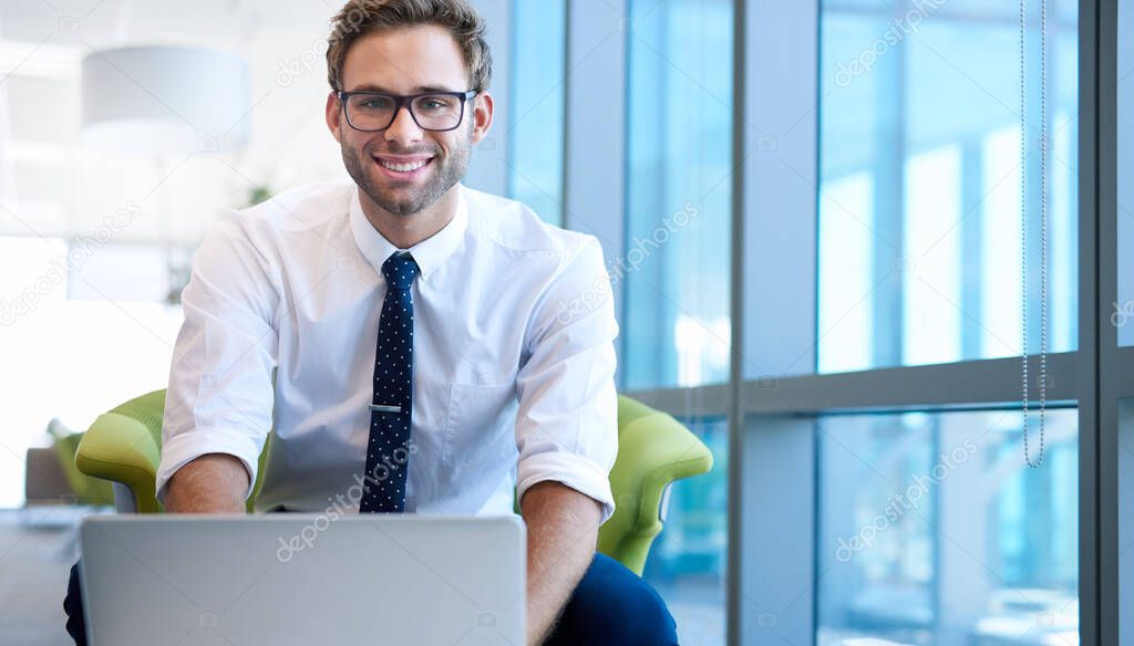 Portrait of a young businessman in a modern office space, typing on his laptop in front of him, and smiling confidently at the camera