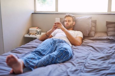 Sabahları yatakta yatarken cep telefonu kullanan pijamalı yakışıklı bir genç.