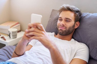 Pijamalı gülümseyen genç adam, sabahları yatakta yatarken cep telefonu kullanıyor.