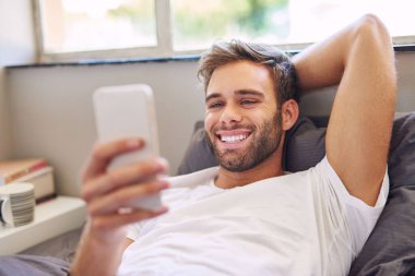 Pijamalı gülümseyen genç adam, sabahları yatakta yatarken cep telefonu kullanıyor.