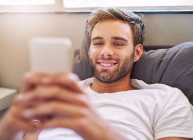 Pijamalı gülümseyen genç adam, sabahları yatakta yatarken cep telefonu kullanıyor.