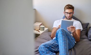 Pijamalı, gözlüklü ve yatakta yatarken dijital tablet kullanan yakışıklı bir genç.