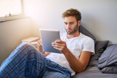 Sabahları yatakta yatarken dijital tablet kullanan pijamalı yakışıklı bir genç.