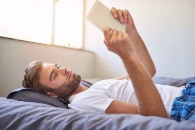 Sabahları yatakta yatarken dijital tablet kullanan pijamalı yakışıklı bir genç.