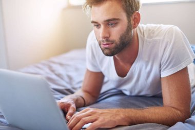 Pijamalı yakışıklı genç adam odaklanmış görünüyor ve sabahları yatakta yatarken dizüstü bilgisayar kullanıyor.