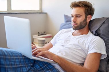 Pijamalı yakışıklı genç adam odaklanmış görünüyor ve sabahları yatakta yatarken dizüstü bilgisayar kullanıyor.