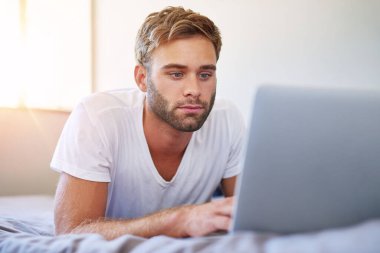 Pijamalı yakışıklı genç adam odaklanmış görünüyor ve sabahları yatakta yatarken dizüstü bilgisayar kullanıyor.
