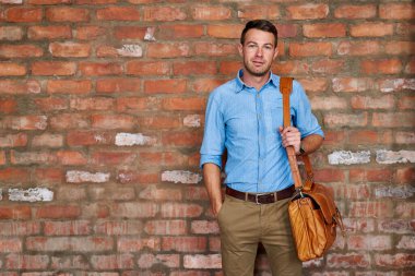 Ofisteki tuğladan bir duvarın önünde çanta taşıyan sıradan giyinmiş genç bir işadamının portresi.