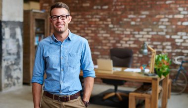 Gözlük takan ve modaya uygun tuğlalı bir ofiste duran sıradan giyinmiş genç bir işadamının portresi.