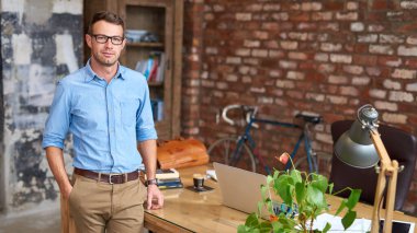 Gözlük takan ve modaya uygun tuğlalı bir ofiste duran sıradan giyinmiş genç bir işadamının portresi.