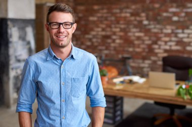 Gözlük takan ve modaya uygun tuğlalı bir ofiste duran sıradan giyinmiş genç bir işadamının portresi.