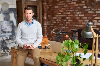 Moda duvarlı bir ofiste masanın üzerinde otururken cep telefonu kullanan sıradan giyinmiş genç bir işadamının portresi.
