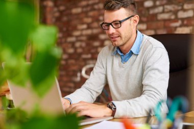 Sıradan giyinmiş genç işadamı, tuğladan bir ofiste masa başında otururken dizüstü bilgisayarda çalışıyor.  