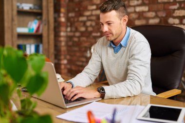 Sıradan giyinmiş genç işadamı, tuğladan bir ofiste masa başında otururken dizüstü bilgisayarda çalışıyor.  