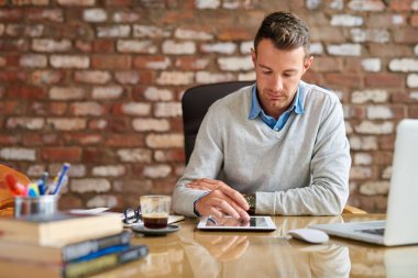 Tuğladan duvarlarla örülü bir ofiste masa başında oturmuş tablet kullanan sıradan giyinmiş genç bir işadamı.  