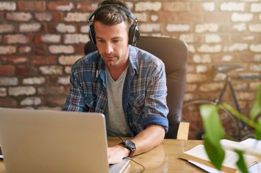 Ofisteki bir masada dizüstü bilgisayarla çalışırken kulaklıkla müzik dinleyen sıradan giyinmiş genç bir tasarımcı.  