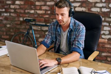 Ofisteki bir masada dizüstü bilgisayarla çalışırken kulaklıkla müzik dinleyen sıradan giyinmiş genç bir tasarımcı.  