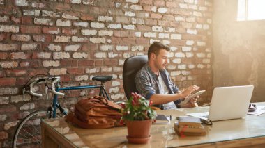 Tuğladan duvarlarla örülü bir ofiste masa başında oturmuş tablet kullanan sıradan giyinmiş genç bir işadamı.  