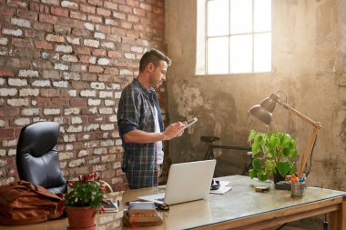 Tuğladan duvarlarla örülü bir ofiste masa başında dururken tablet kullanan sıradan giyinmiş genç bir tasarımcı.  