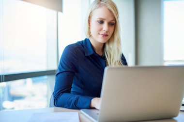 Genç bir iş kadını, parlak, modern bir ofiste masasında tek başına otururken dizüstü bilgisayarda çalışıyor.