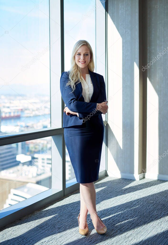 Portrait of a young businesswoman smiling confidently while standing with her arms crossed by windows in a modern office building overlooking the city