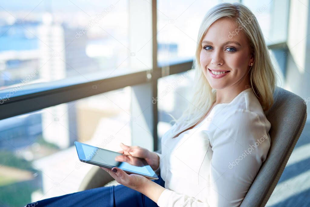 Portrait of a smiling young businesswoman using a digital tablet while sitting in a chair by windows in a modern office building overlooking the city 