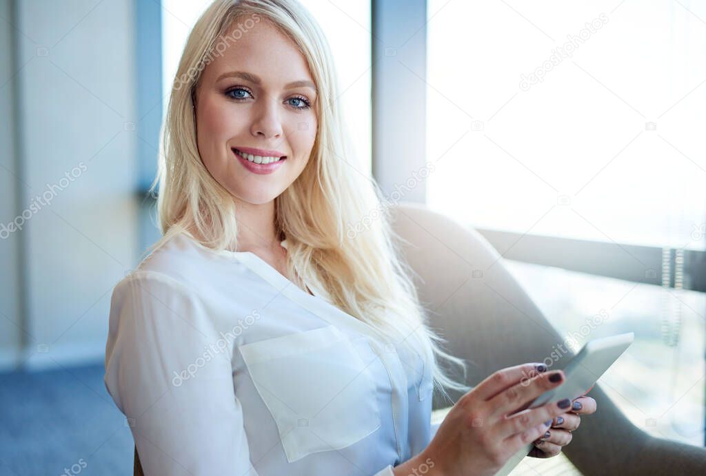 Portrait of a smiling young businesswoman sitting in a chair by windows in a modern office working online with a digital tablet 
