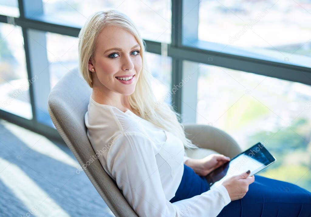 Portrait of a smiling young businesswoman working with a digital tablet while sitting in a chair by windows in a modern office building 