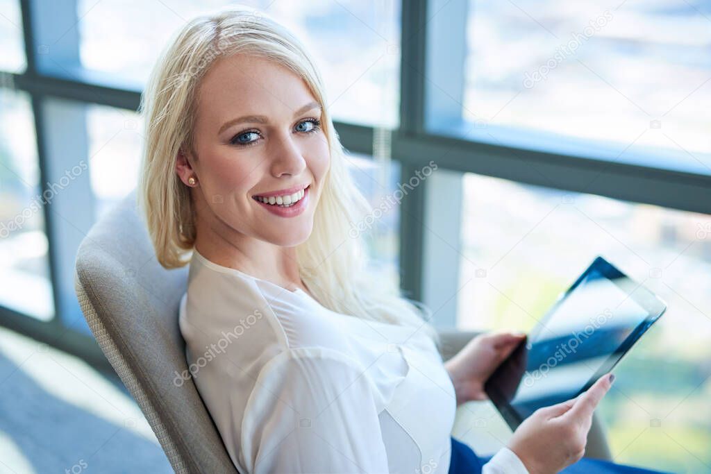 Portrait of a smiling young businesswoman working on a digital tablet while sitting in a chair by windows in a modern office building 
