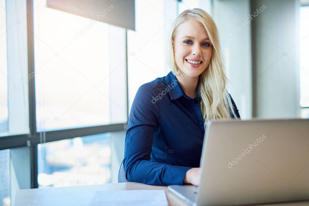 Portrait of a smiling young businesswoman sitting alone at a desk in a modern office working on a laptop   