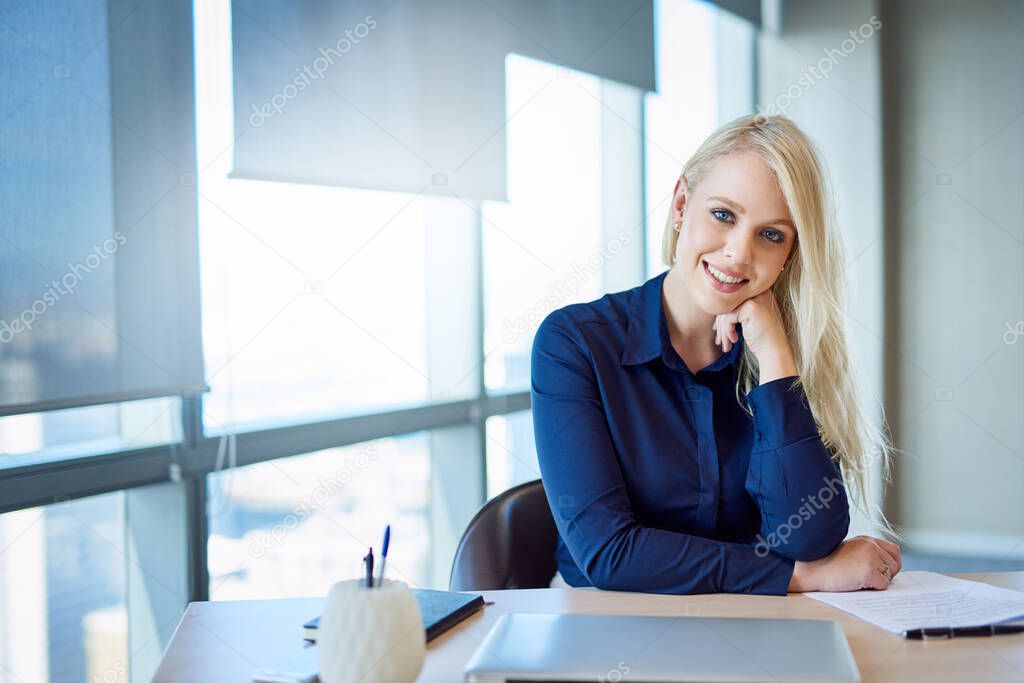 Portrait of a smiling young businesswoman sitting at her desk in an office with her hand on her chin 
