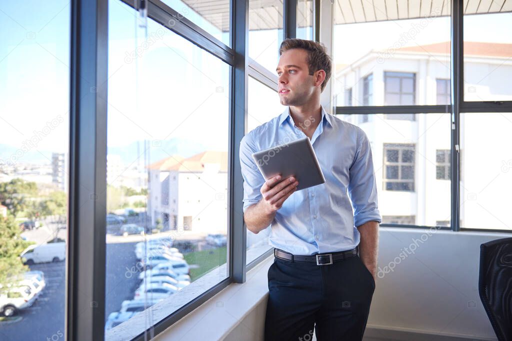 Handsome young businessman, standing in his office with his hand in his pocket and holding a digital tablet, while looking out of his office window and thinking