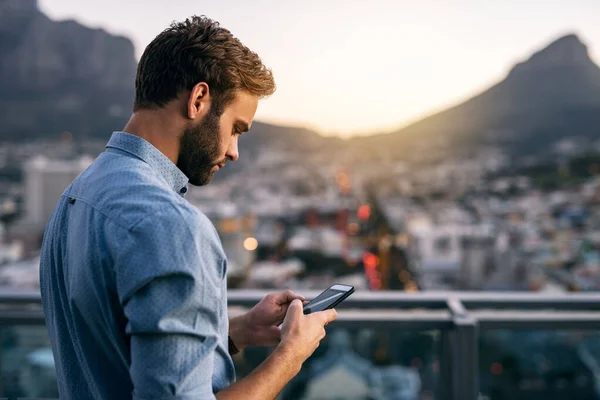 Focused young businessman sending texts on a cellphone while standing on an office building balcony overlooking the city at dusk