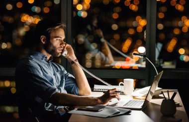 Genç işadamı telefonda konuşuyor ve gece geç saatlerde ofisteki masasında dizüstü bilgisayarını kullanarak şehre bakan pencerelerin önünde. 