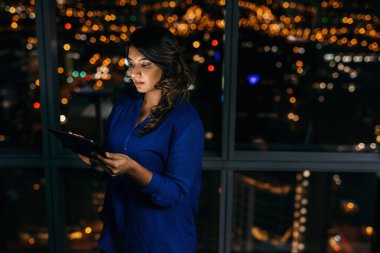 Dijital tablet kullanan genç bir iş kadını gece geç saatlerde bir ofiste tek başına şehre tepeden bakan pencerelerin önünde dikiliyor. 