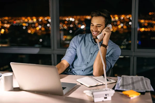 Smiling young businessman talking on the phone while working overtime at his office desk late into the night in front of windows overlooking the city