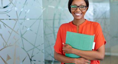 Modern bir ofiste dururken elinde dosyalar olan ve gözlük takan gülümseyen bir ofis çalışanının portresi.