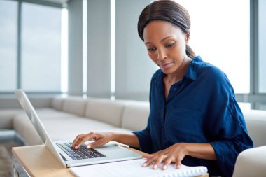 Odaklanmış genç iş kadını notlar okuyor ve modern bir ofiste masada otururken bir dizüstü bilgisayar üzerinde çalışıyor.