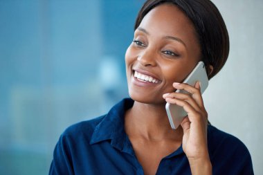 Modern bir ofiste pencerelerin önünde dururken cep telefonuyla gülümseyen ve konuşan çekici genç bir iş kadını.