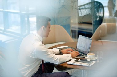 Odaklanmış genç işadamı bir ofisteki bir odada tek başına oturmuş dizüstü bilgisayarla çalışıyor.