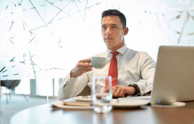 Beyaz gömlekli ve kırmızı kravatlı genç bir işadamı ofisteki bir masada oturmuş laptopla çalışırken kahve içiyor.