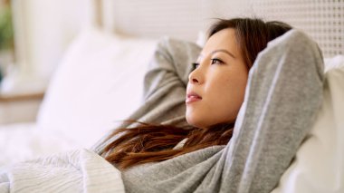 Genç Asyalı kadın sabahın erken saatlerinde yatağında elleri başının arkasında yatıyor.