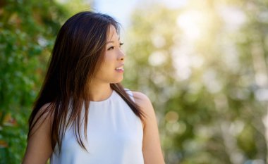 Gülümseyen Asyalı bir kadın güneşli bir günde parkta tek başına yürüyüşe çıkıyor.