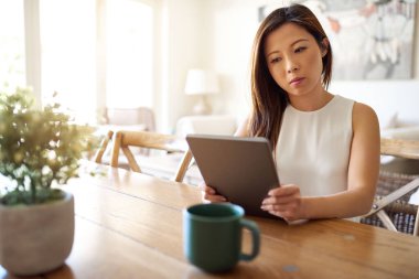 Çekici genç Asyalı kadın elinde tabletle çalışıyor ve evde tek başına oturmuş kahve içiyor.