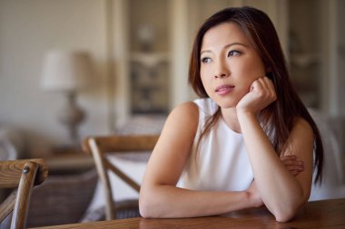 Genç Asyalı kadın eli çenesinde bir masada otururken derin düşüncelere dalmış.